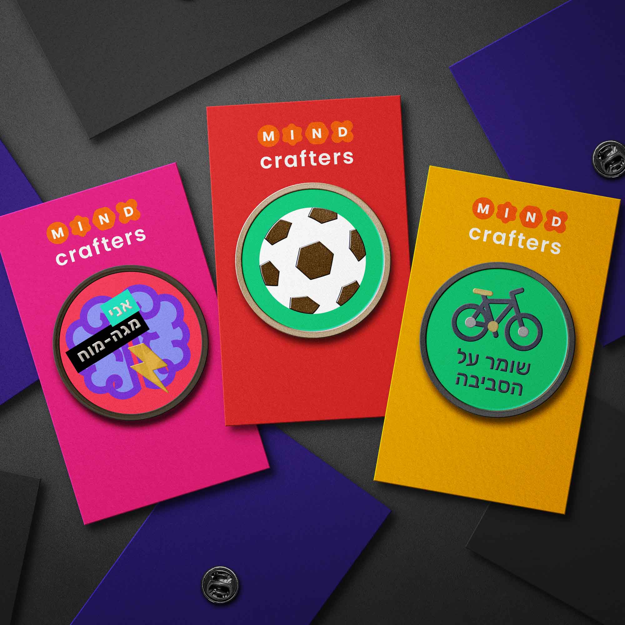 שלושה תגי מתכת צבעוניים מבית ספר MindCrafters – תג ורוד עם ציור מוח וברק וכיתוב "אנחנו מגה-מוח", תג אדום עם כדורגל, ותג צהוב עם אופניים וכיתוב "שומר על הסביבה". עיצוב גרפי צבעוני ומודרני שמייצג תחומי עניין שונים של תלמידים – חשיבה, ספורט ואחריות סביבתית.